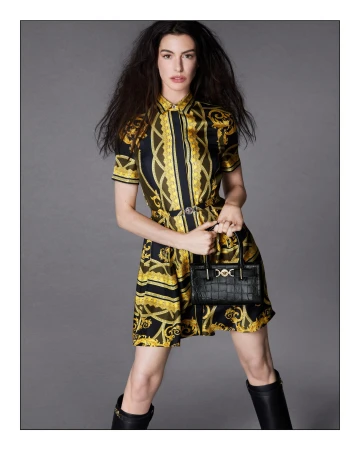 Anne Hathaway wearing a printed yellow and black dress holding a black handbag against a grey backdrop