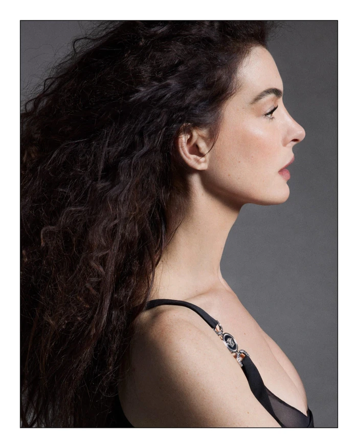 Profile portrait of actress Anne Hathaway with long dark curly hair wearing a black dress for Versace