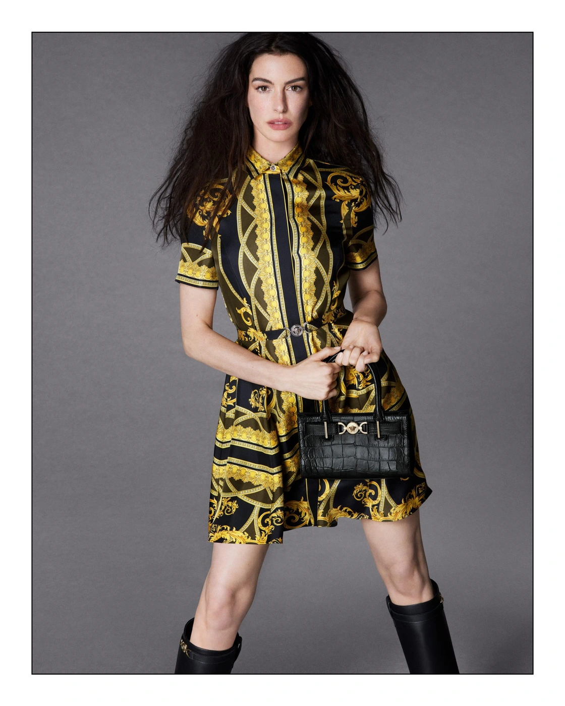 Anne Hathaway wearing a printed yellow and black dress holding a black handbag against a grey backdrop