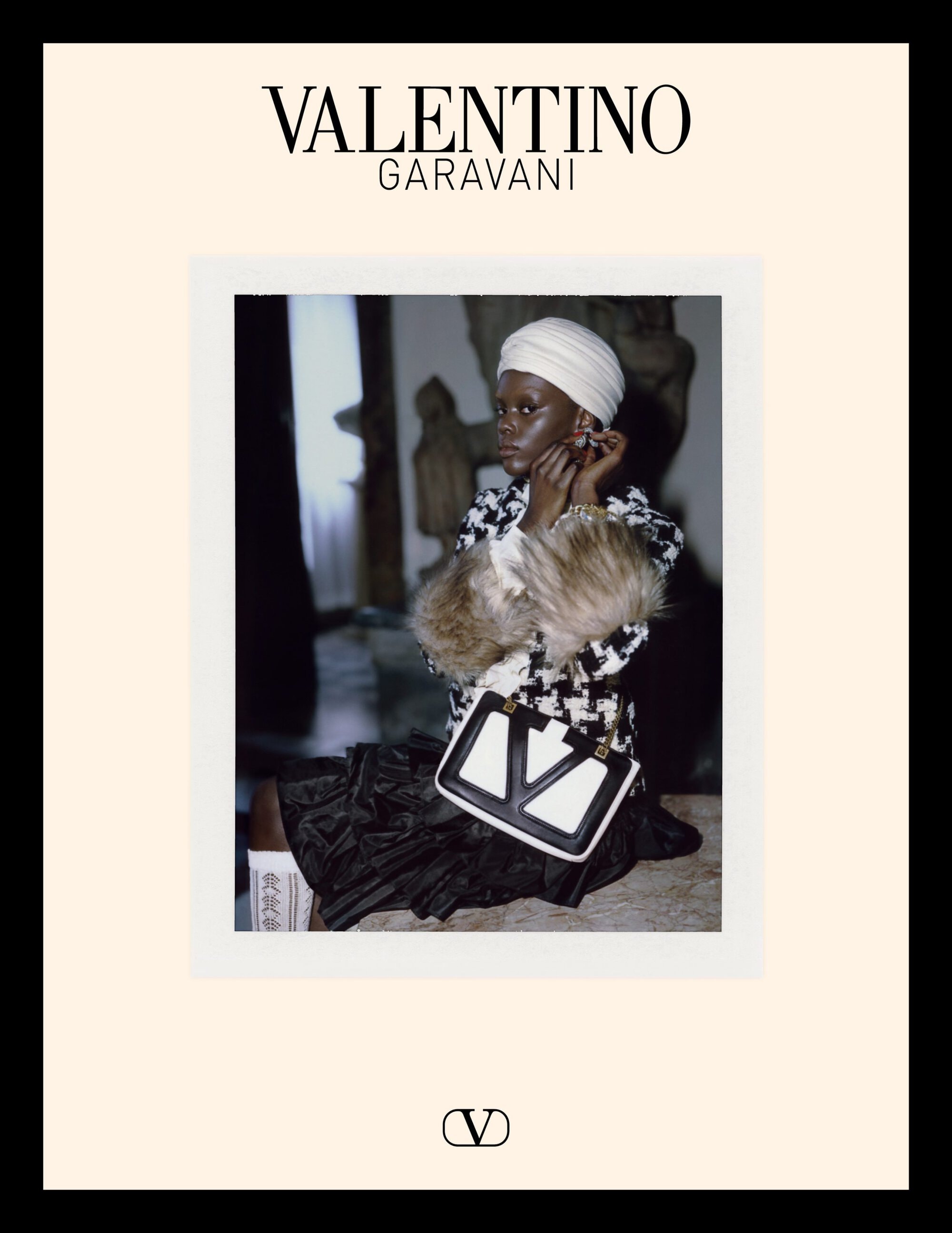 Model wearing a houndstooth jacket and white turban holding a structured black and white handbag against an interior backdrop