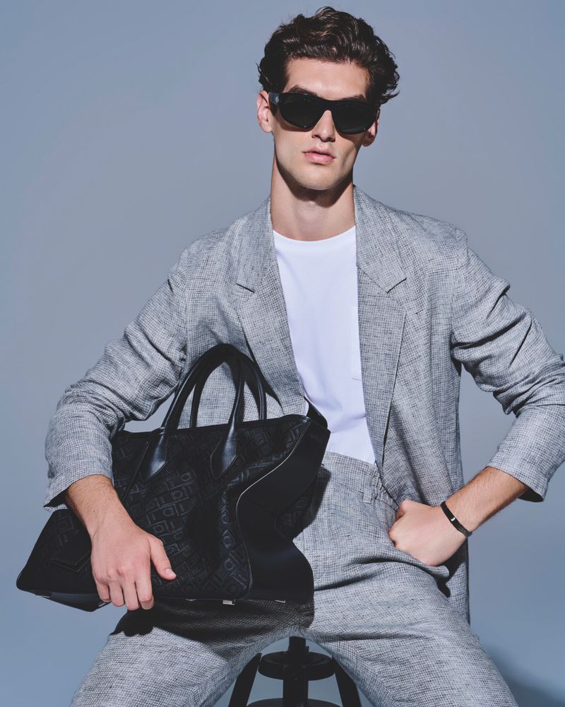 Adult male model in a grey suit and sunglasses seated with a black patterned tote bag in a studio with grey backdrop and neutral tones.