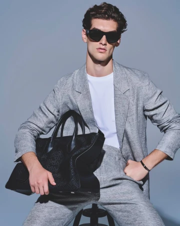 Adult male model in a grey suit and sunglasses seated with a black patterned tote bag in a studio with grey backdrop and neutral tones.