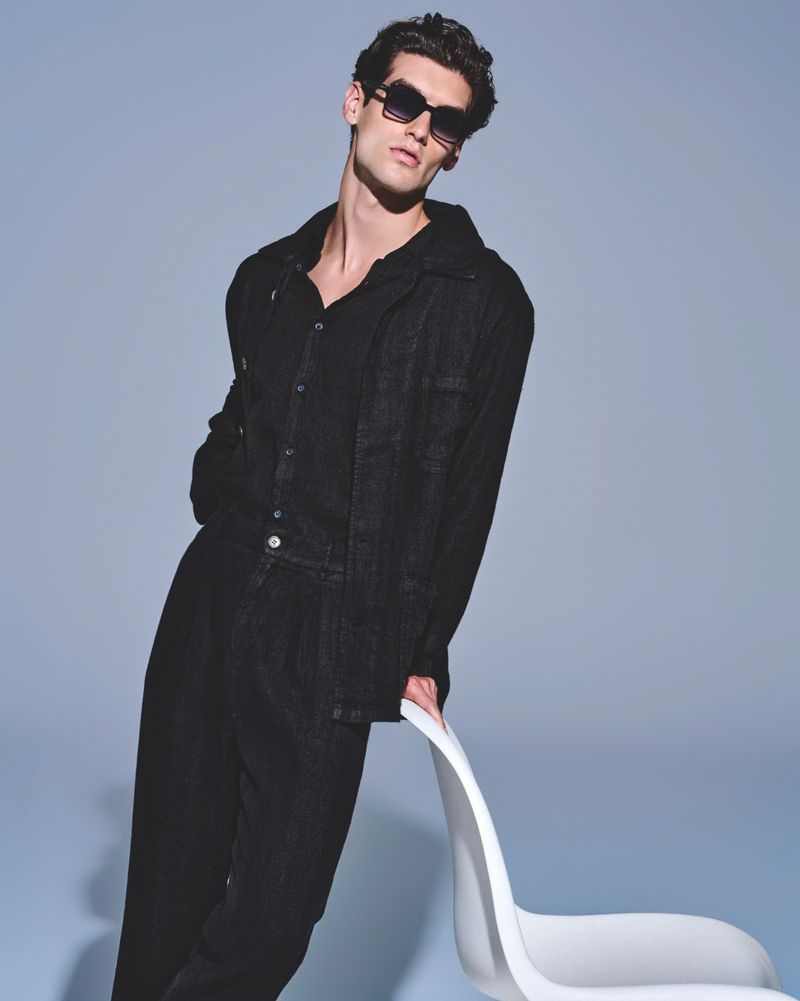 Studio shot of a dark-haired male fashion model in a black denim suit and sunglasses, leaning on a white chair against a blue background.