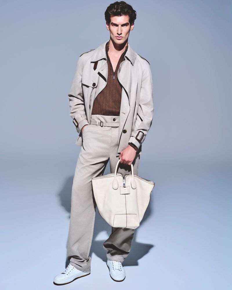Full shot of a male fashion model posing in a trench coat, brown shirt, beige pants, and white sneakers while holding a white tote bag.