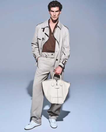 Full shot of a male fashion model posing in a trench coat, brown shirt, beige pants, and white sneakers while holding a white tote bag.