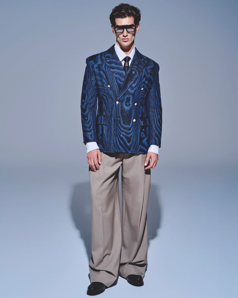 Man in blue patterned double-breasted blazer, white shirt, and beige wide-leg pants posing in a studio with a neutral background.