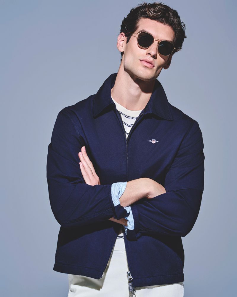 A fashionable adult male poses indoors wearing a navy jacket, white pants, striped shirt, and sunglasses against a grey background.