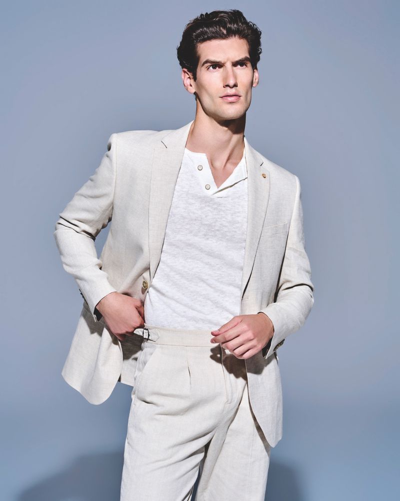 Man in a beige suit and white shirt poses against a grey background, looking away from camera. Fashion and portrait photography.