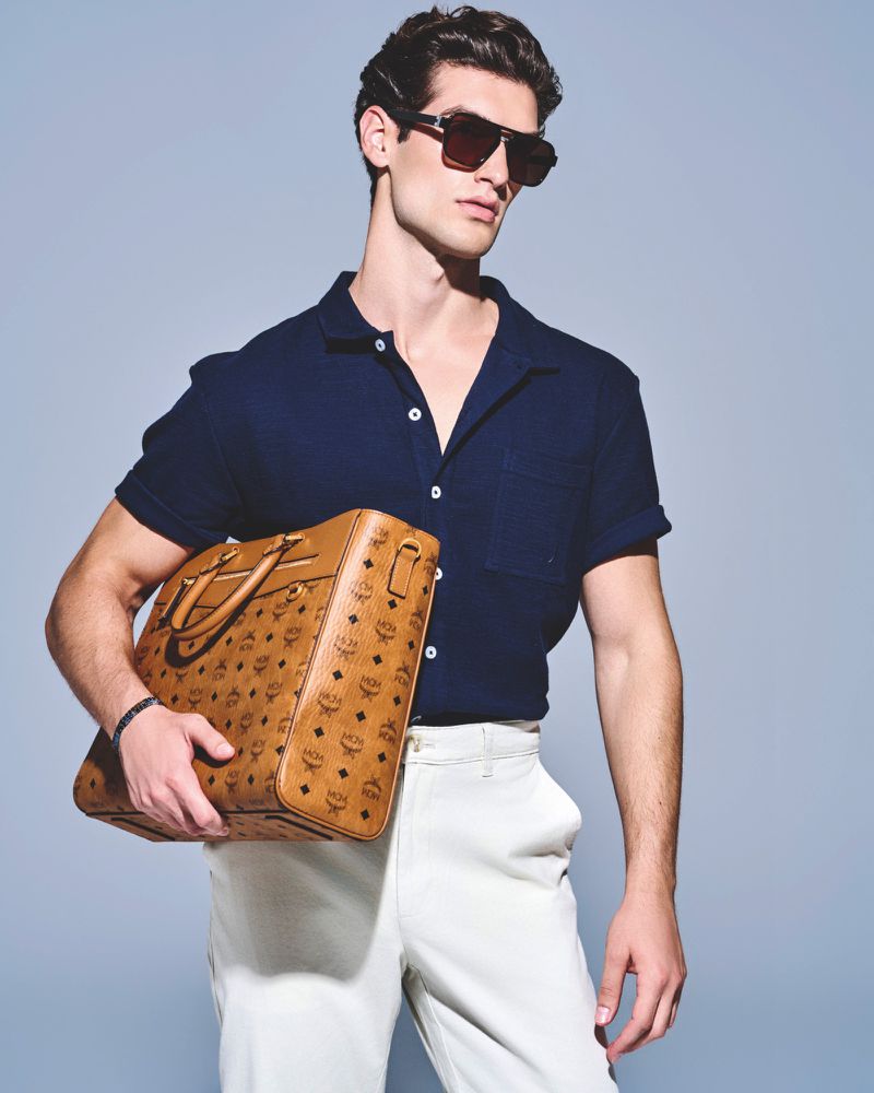 A brunette adult male poses in a navy short-sleeved shirt and white slacks, holding a brown MCM logo-patterned briefcase, indoors.