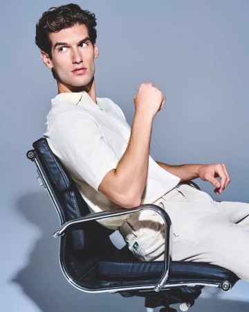Portrait of a dark-haired adult male sitting in a black office chair, wearing a collared shirt and light-colored pants against a gray backdrop.