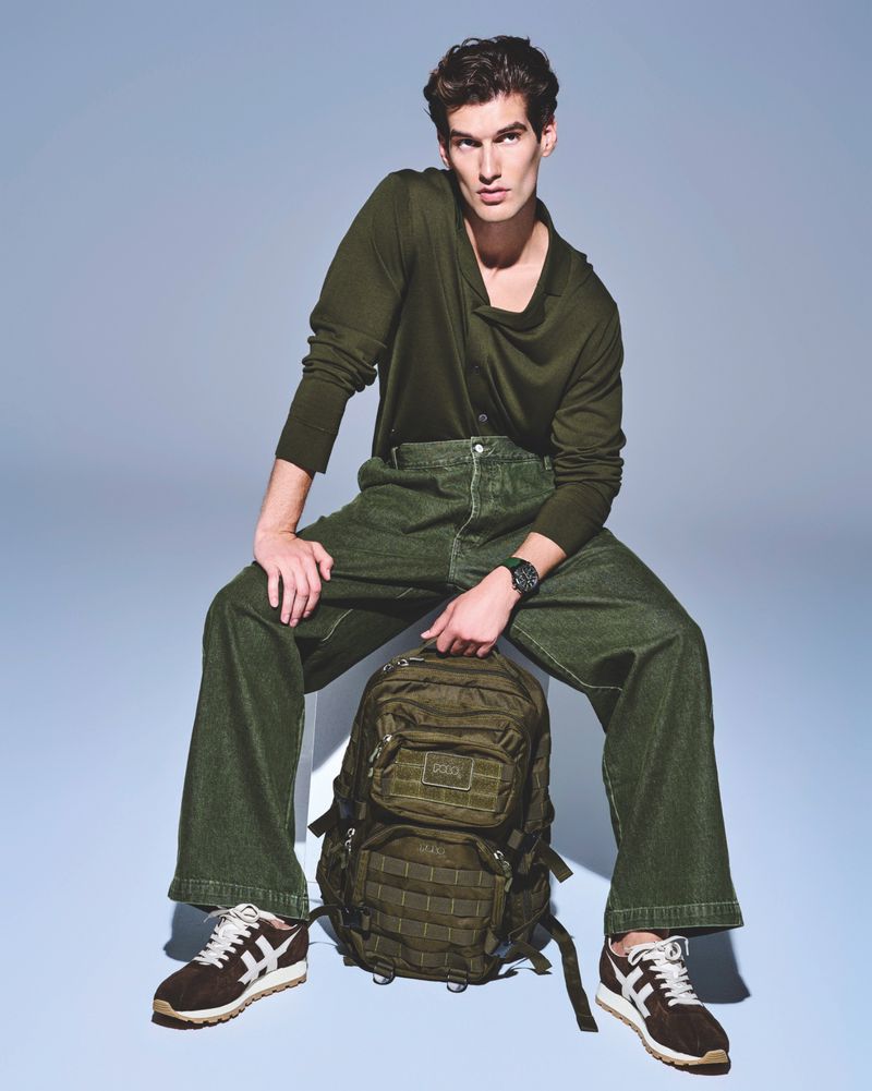 Man in olive-green casual outfit poses sitting with backpack and sneakers in a studio setting, providing fashion-forward visual appeal.