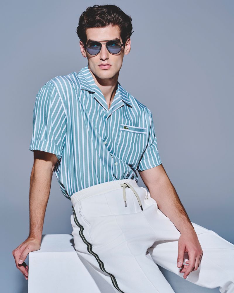 A young adult male is posing in a fashion shot, wearing a striped shirt and white pants, set against a neutral background in a photo studio.