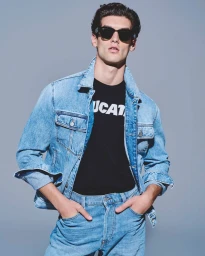 A man wearing a light blue denim jacket, dark sunglasses, and black t-shirt against a plain grey backdrop for Esquire Greece.