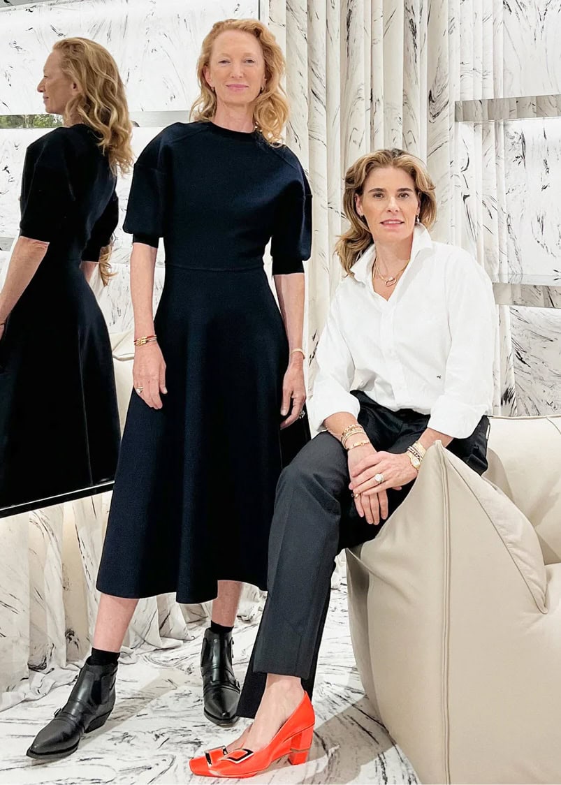 Two women posing in an interior space, one standing in a black dress and one seated wearing a white shirt and dark trousers