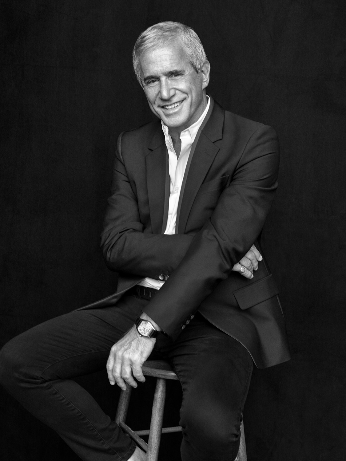Black and white portrait of a smiling older man seated on a stool wearing a dark suit jacket and white shirt.