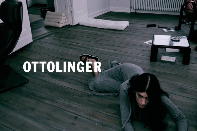 Woman in grey long-sleeve dress lying on a dark wooden floor inside a room