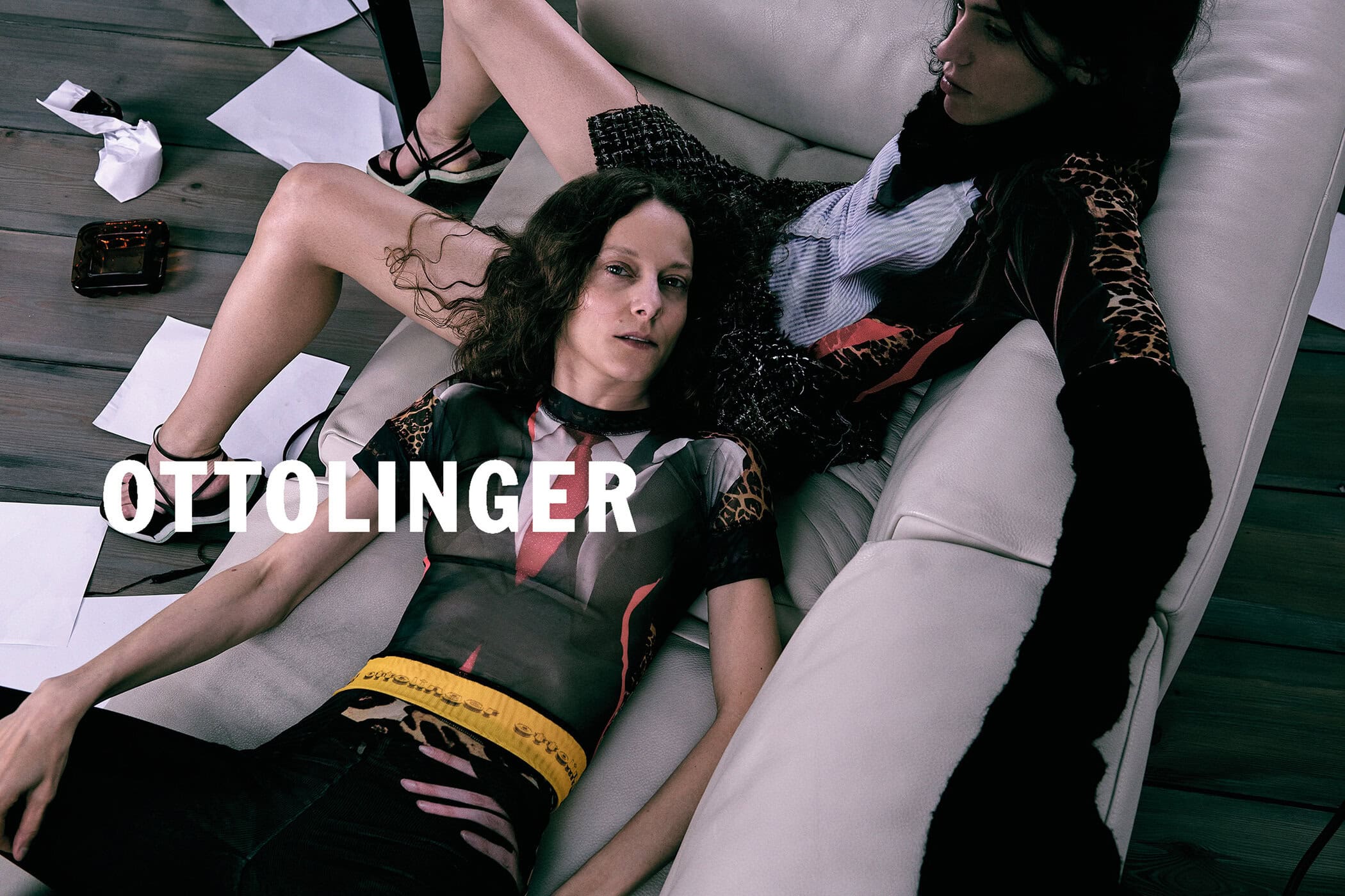 Two models reclining on a white leather lounge chair with scattered papers on a dark wooden floor.