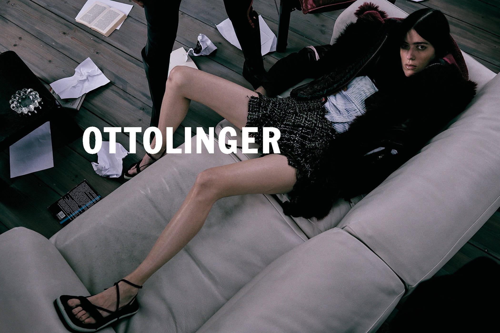 Model lying on a pale leather sofa wearing a tweed skirt and dark jacket, surrounded by scattered papers.