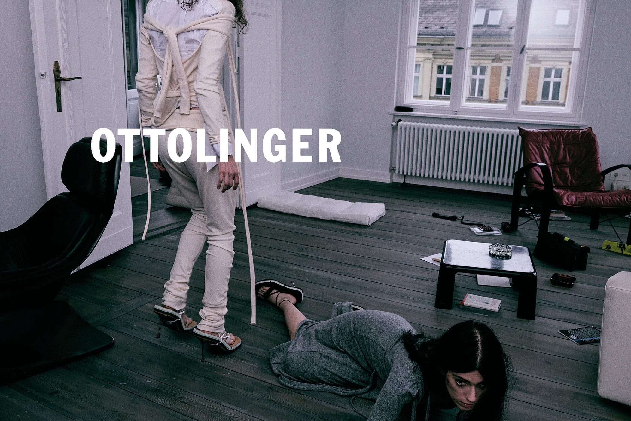 Model wearing cream strapped top and trousers standing over another model lying on a wooden floor in a room