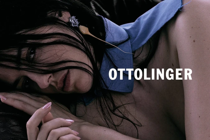 Close-up portrait of a woman with dark hair and blue collared shirt resting her head on her hand with the word OTTOLINGER overlaid.