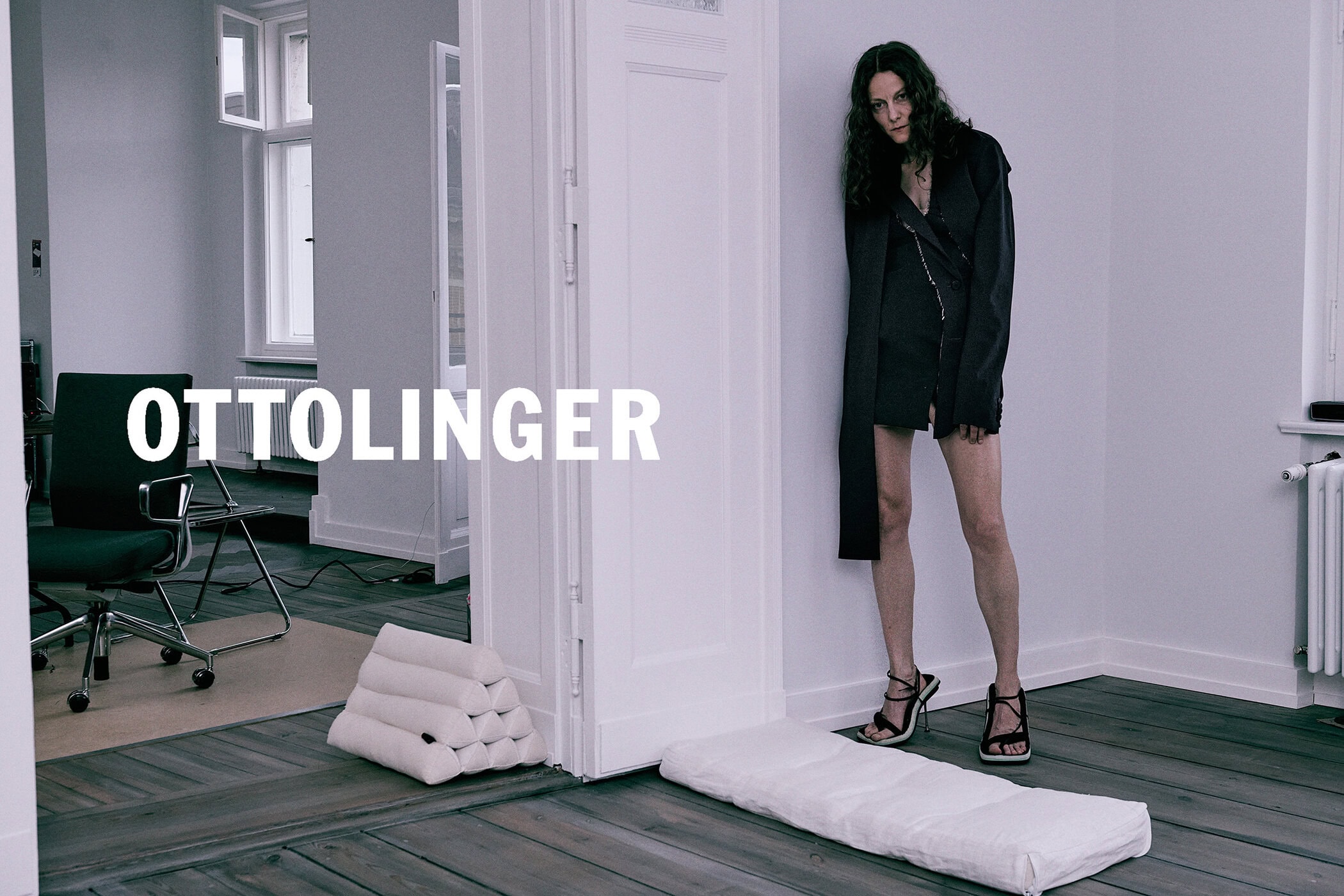 Woman standing in a minimalist room leaning against a white doorframe wearing a dark asymmetric coat and strappy heels
