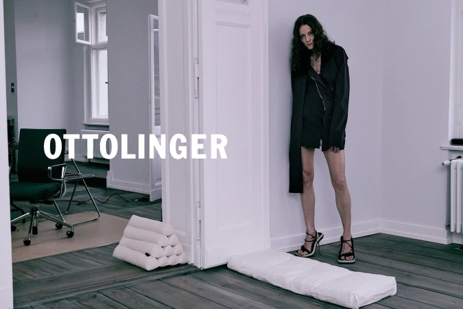 Woman standing in a minimalist room leaning against a white doorframe wearing a dark asymmetric coat and strappy heels