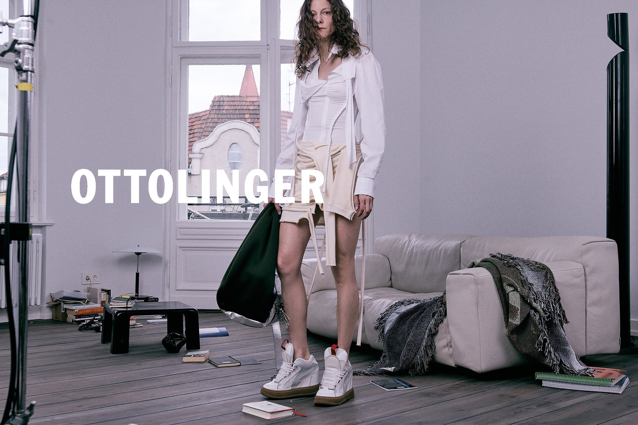 Model wearing a white shirt and cream skirt holding a green bag inside a room with a white sofa and books on the floor