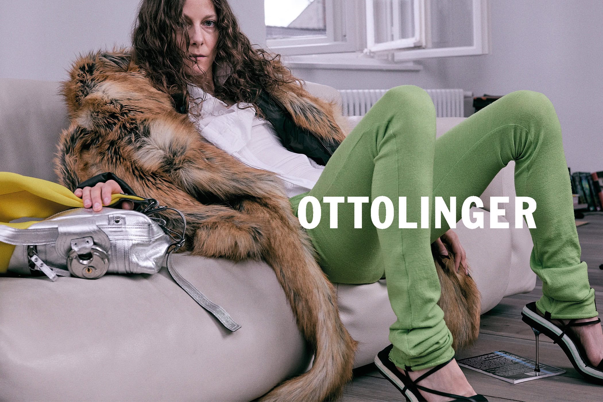 Model with long dark curly hair wearing green trousers and faux fur coat lounging on a sofa, with silver bag.