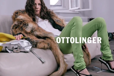 Model with long dark curly hair wearing green trousers and faux fur coat lounging on a sofa, with silver bag.