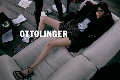 Model lying on a pale leather sofa wearing a tweed skirt and dark jacket, surrounded by scattered papers.