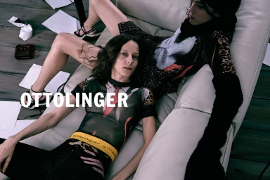 Two models reclining on a white leather lounge chair with scattered papers on a dark wooden floor.