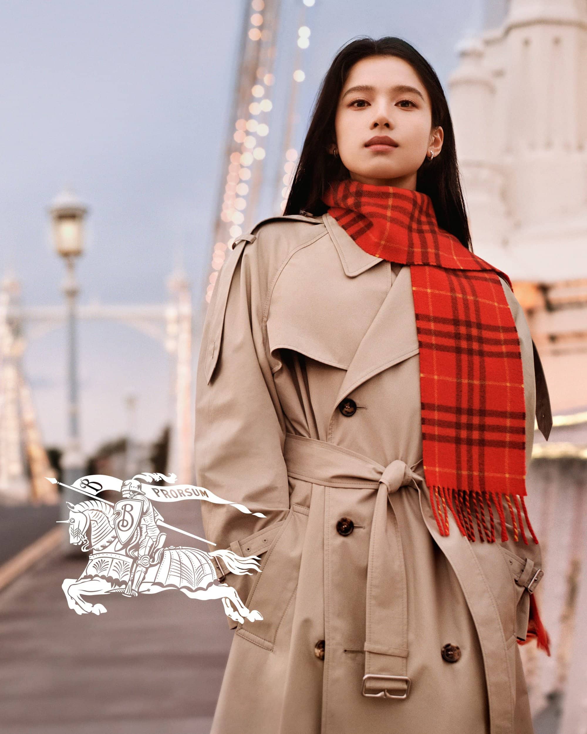 Zhang Jingyi wearing a beige trench coat and red plaid scarf against an outdoor bridge background