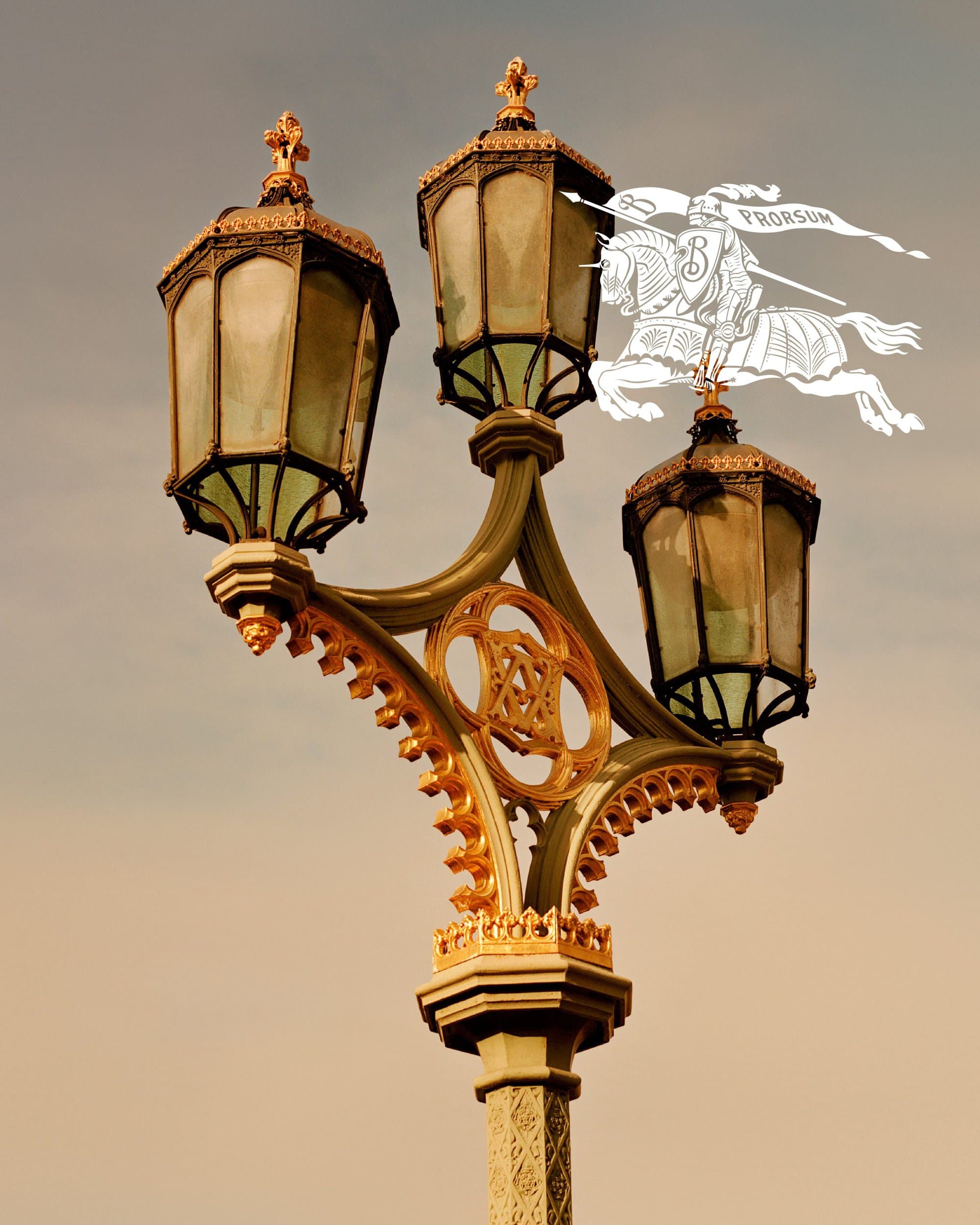 An ornate triple-lamp street post with gold detailing against a muted sky, featuring the Burberry Prorsum knight logo.