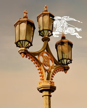 An ornate triple-lamp street post with gold detailing against a muted sky, featuring the Burberry Prorsum knight logo.