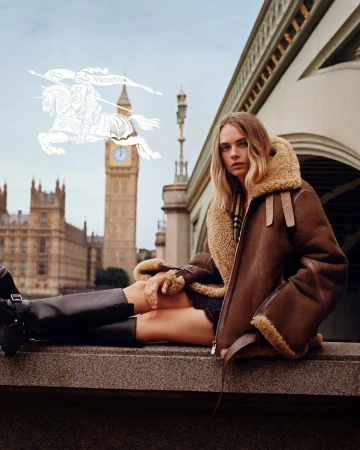 Cara Delevingne seated on a stone ledge wearing a brown shearling jacket and black boots in London.
