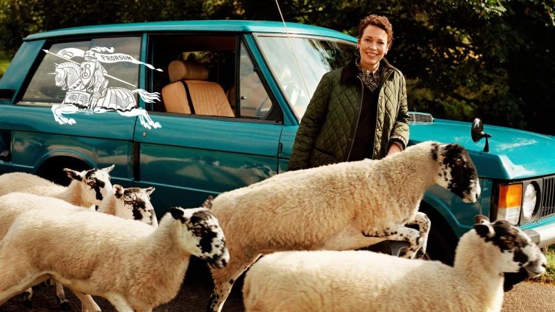 Olivia Colman wearing a quilted green jacket standing beside a teal car with sheep moving in the foreground