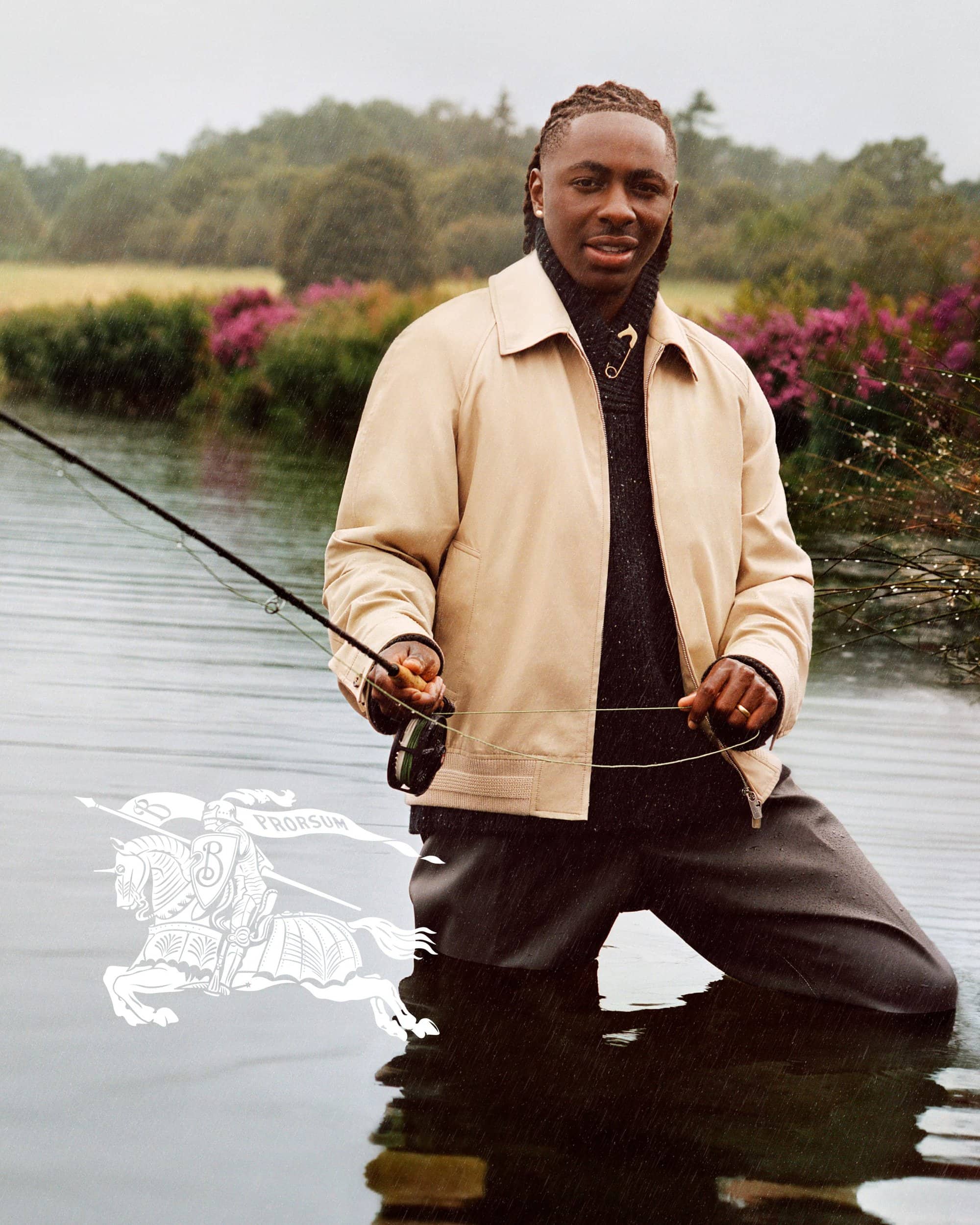 Eberechi Eze wading in a river holding a fishing rod for Burberry wearing a beige jacket and dark sweater