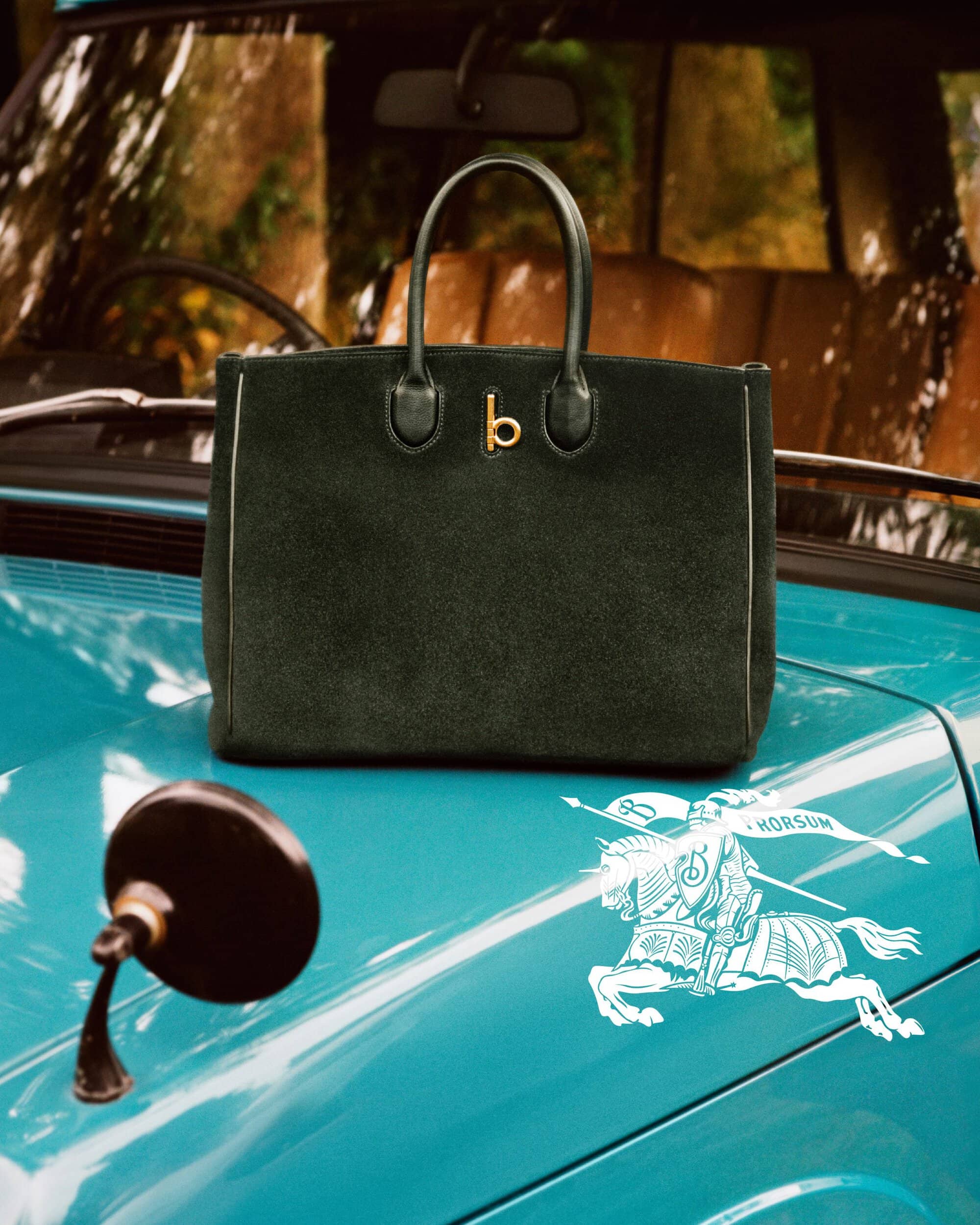 Dark green suede handbag resting on the teal hood of a classic car with a white equestrian knight logo.
