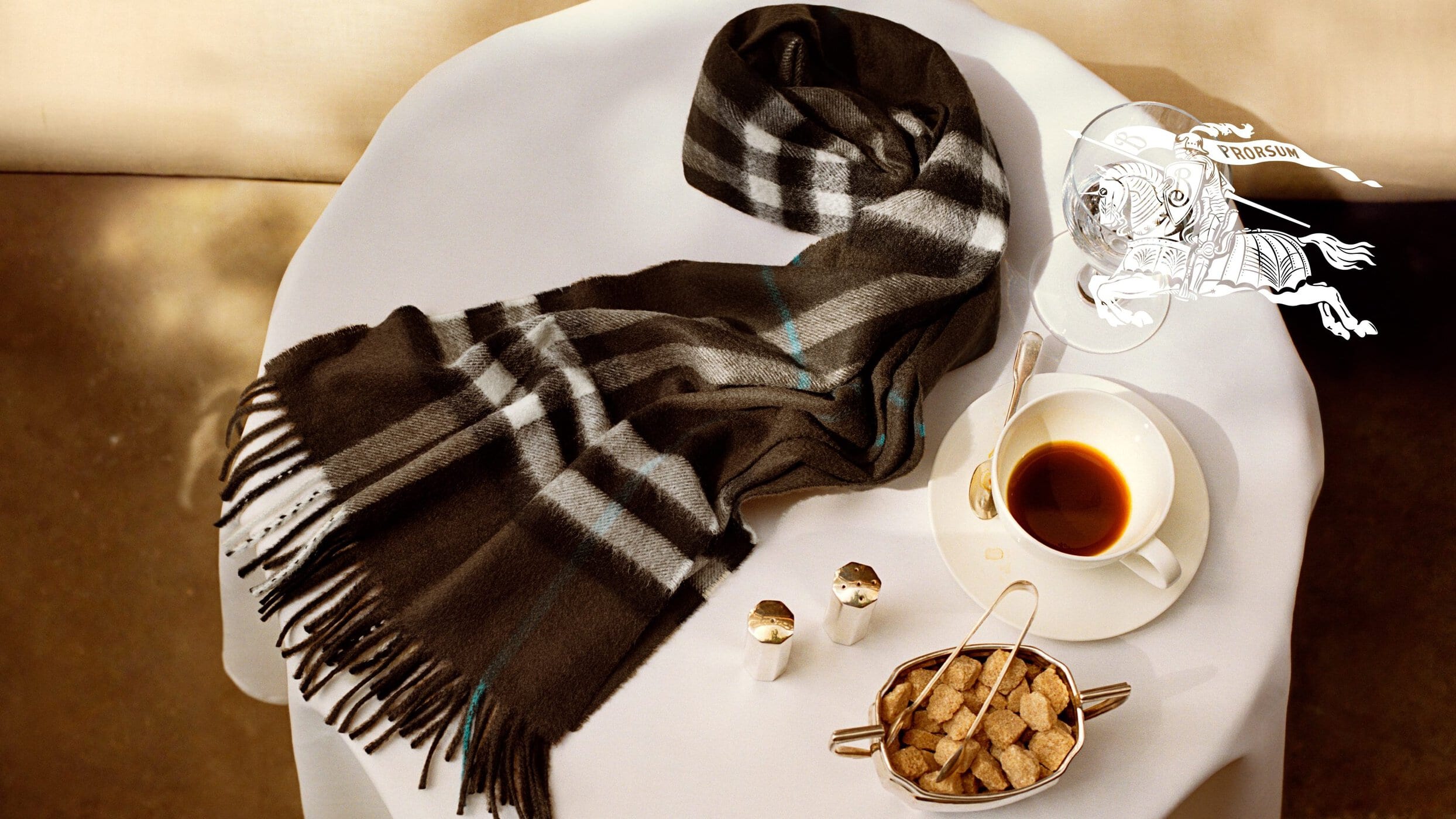 Burberry plaid scarf resting on a small white table with a cup of coffee and sugar cubes