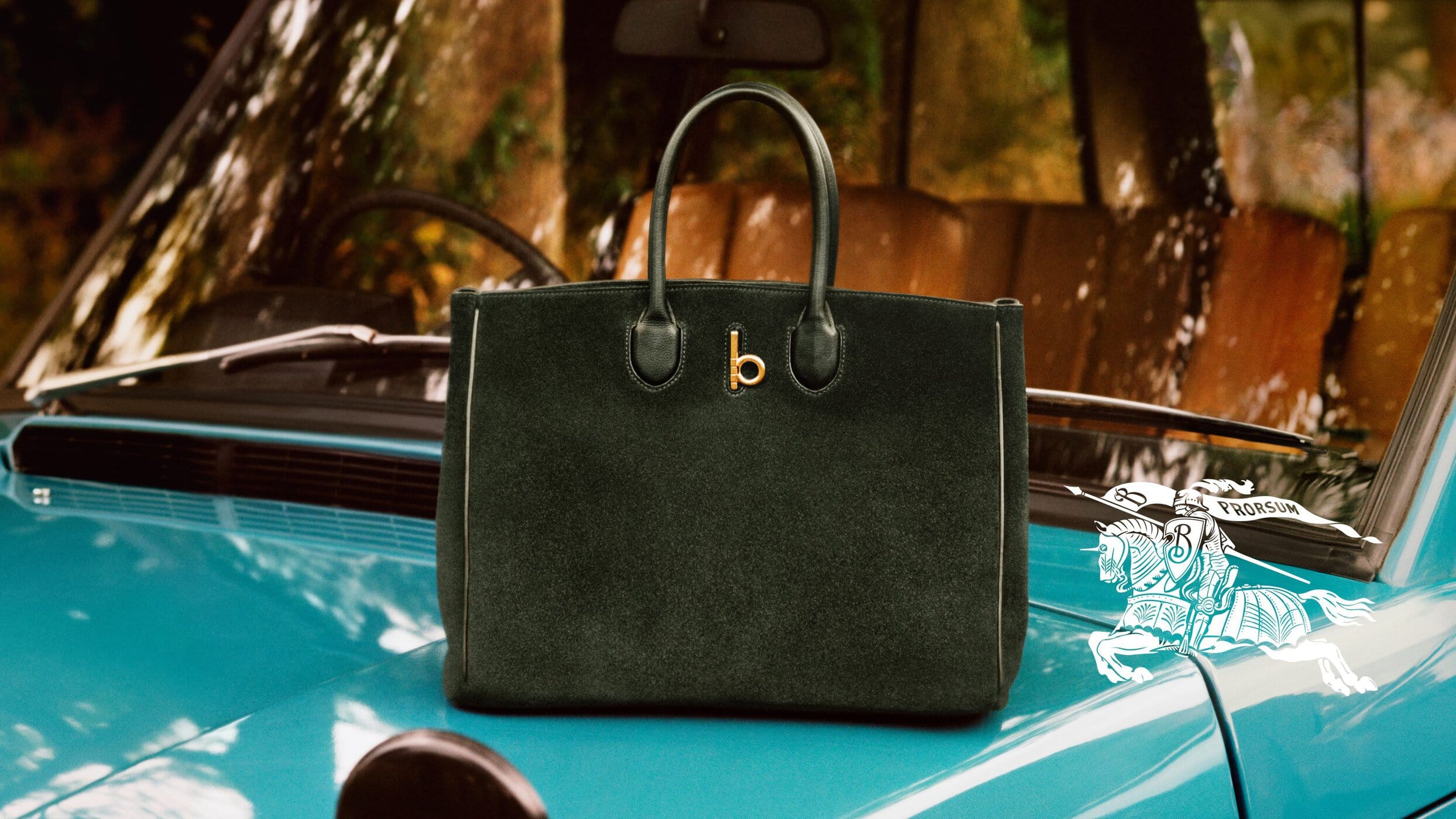 Dark green suede handbag resting on the turquoise blue hood of a vintage car with Burberry branding.
