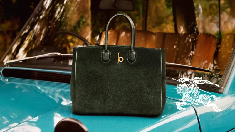 Dark green suede handbag resting on the turquoise blue hood of a vintage car with Burberry branding.