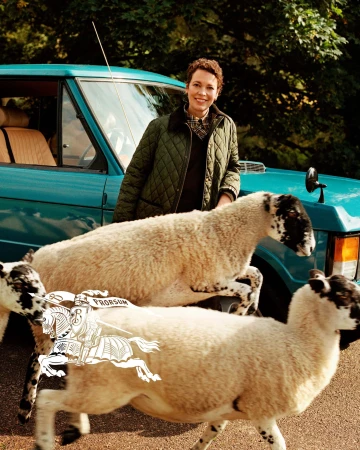 Olivia Colman wearing a quilted green jacket standing beside a blue car with sheep in the foreground