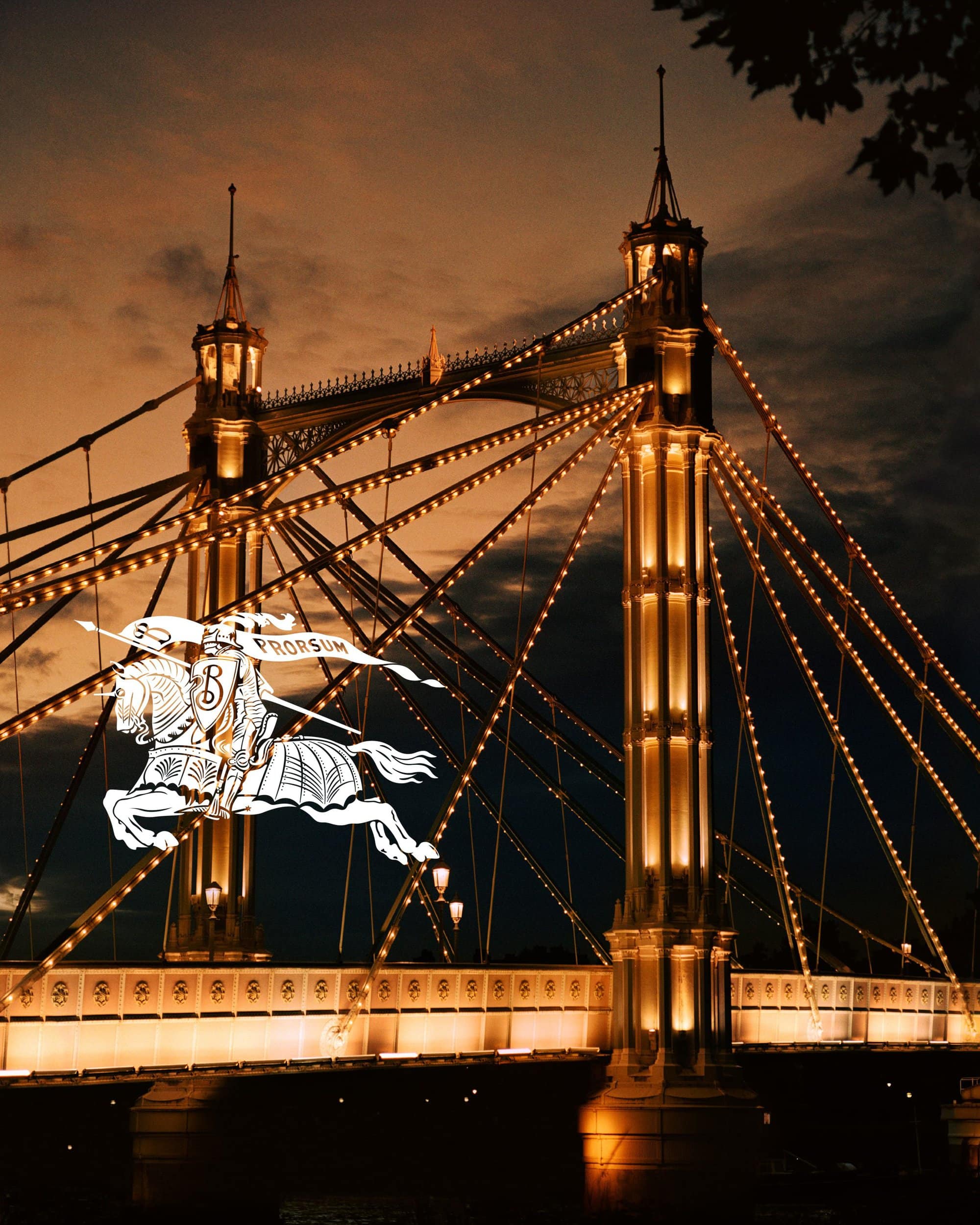Illuminated suspension bridge at night with warm lighting and a prominent white Burberry equestrian knight logo overlaid on the left side.
