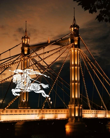 Illuminated suspension bridge at night with warm lighting and a prominent white Burberry equestrian knight logo overlaid on the left side.
