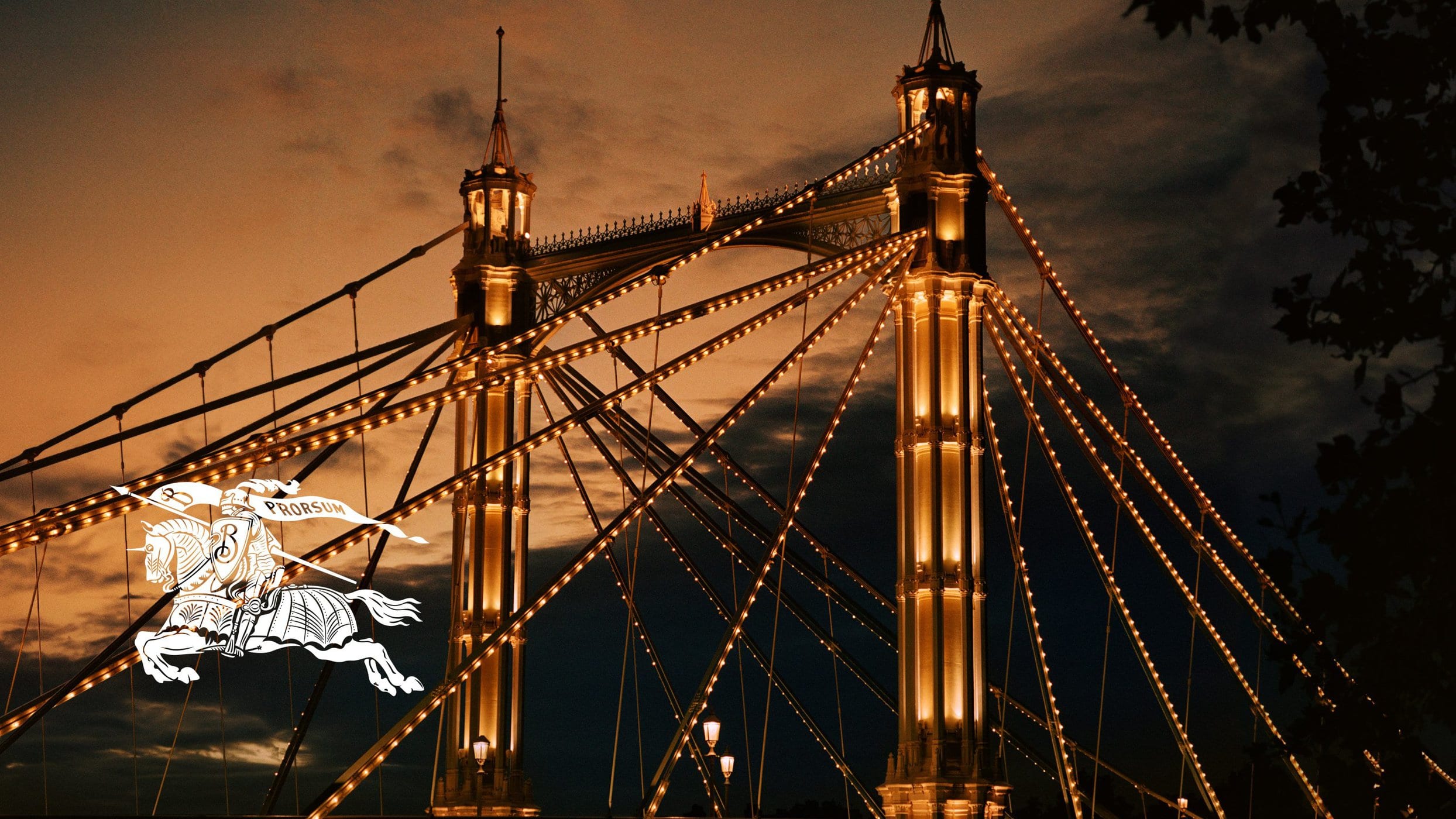 Illuminated suspension bridge towers at dusk with warm string lights outlining the cables and structural elements.