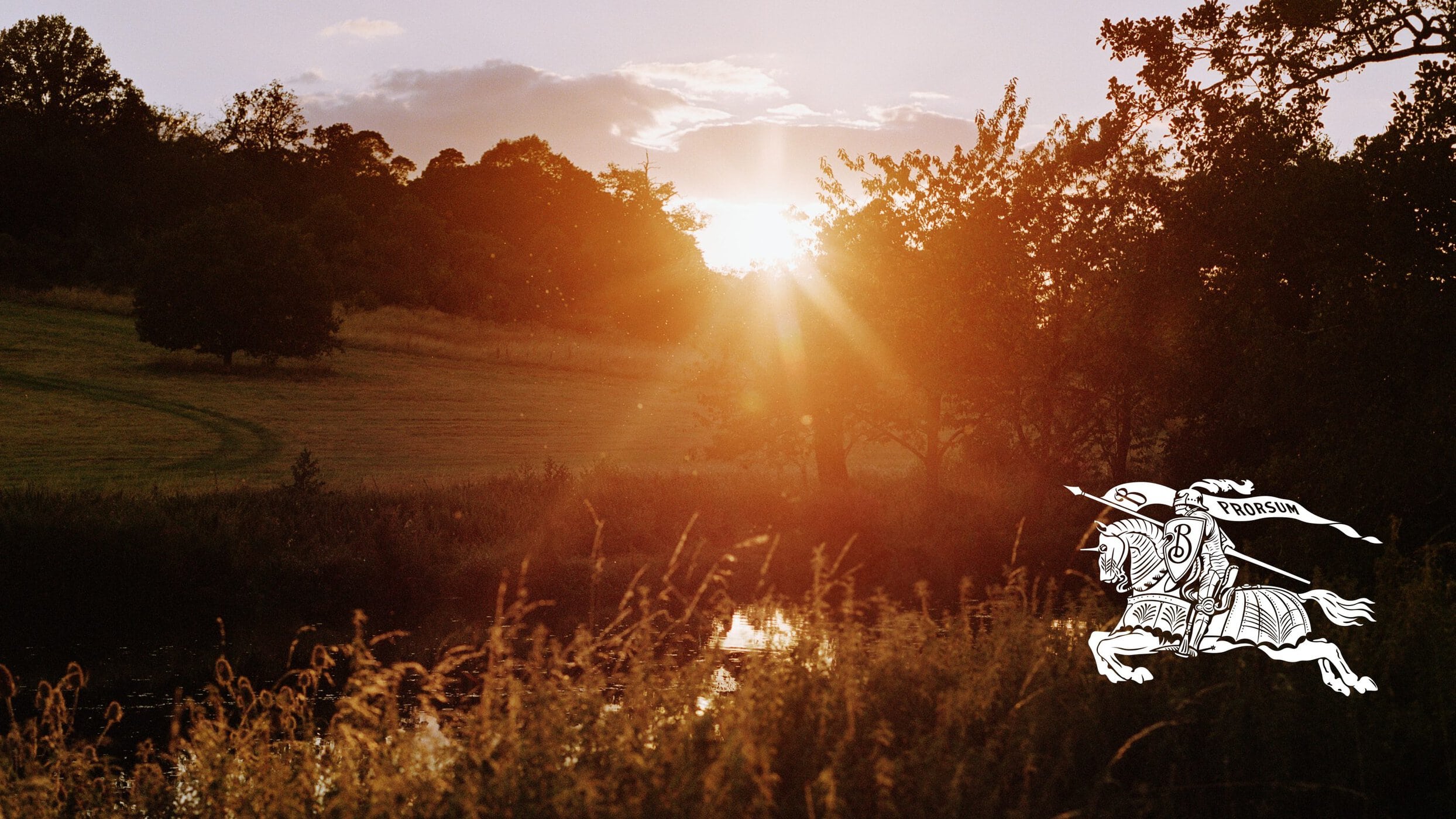 Golden sunset sky over a grassy field with trees and a river, featuring the white Burberry equestrian knight logo in the bottom right.