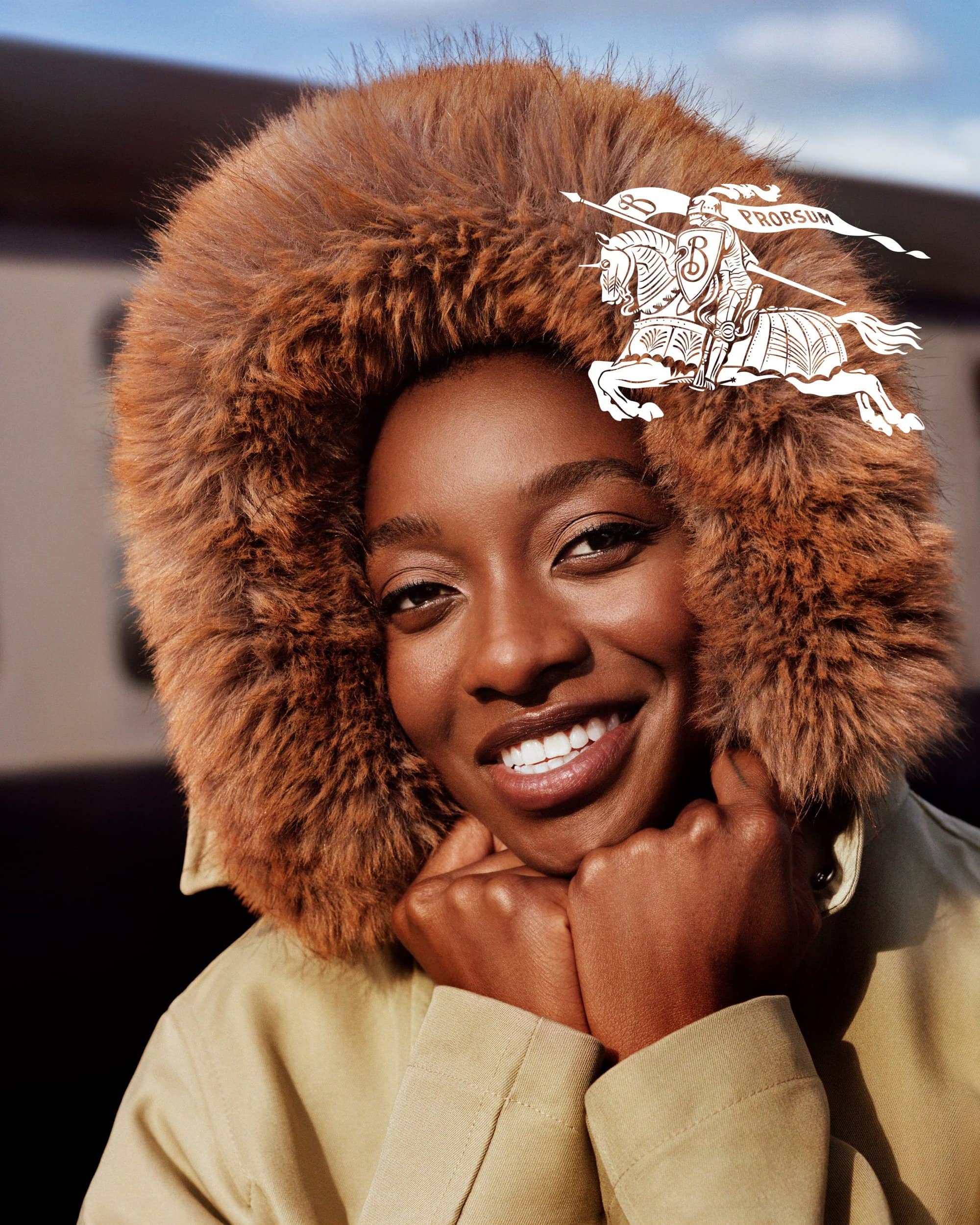 Portrait of a Black woman wearing a brown furry hood and a tan jacket, smiling with hands under chin, for Burberry.