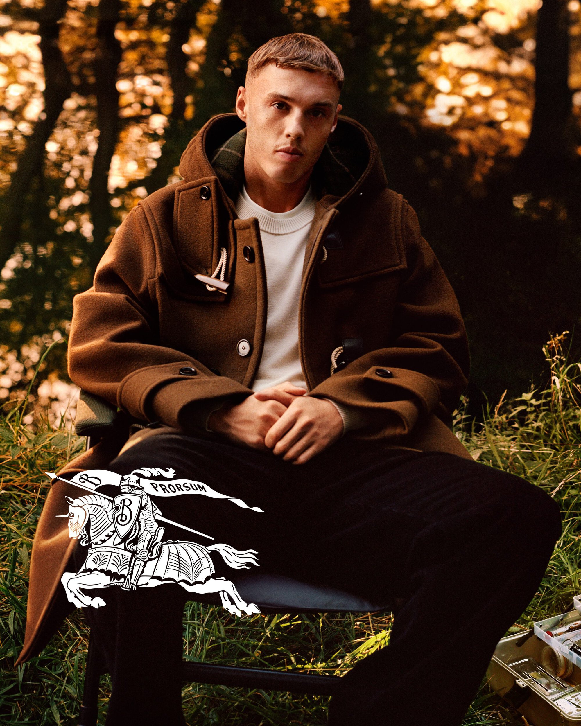 Cole Palmer wearing a brown duffle coat and white t-shirt seated outdoors in a woodland setting for Burberry