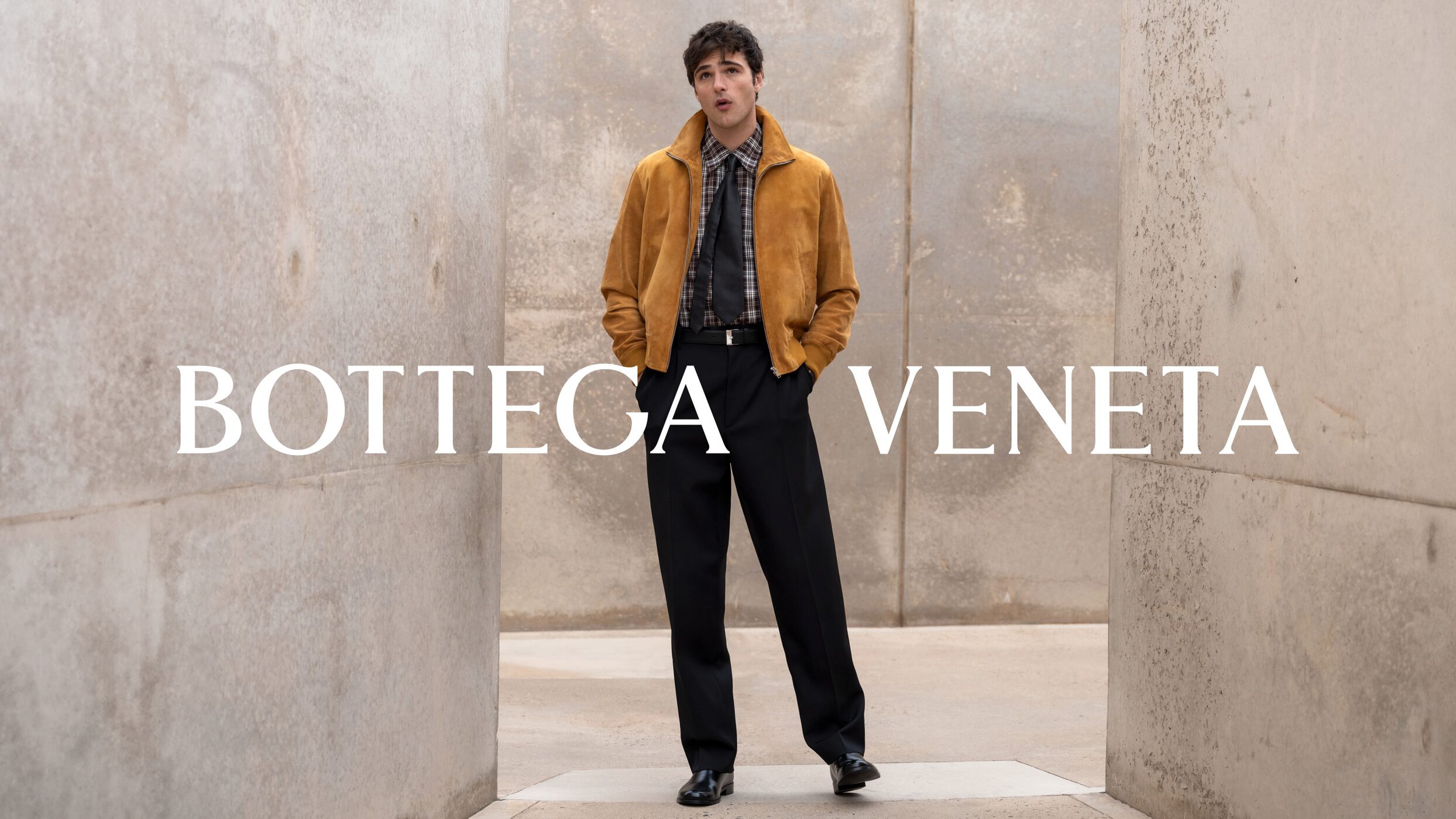 Jacob Elordi wearing a yellow suede jacket and plaid shirt standing between concrete walls for Bottega Veneta Fall 2024 campaign