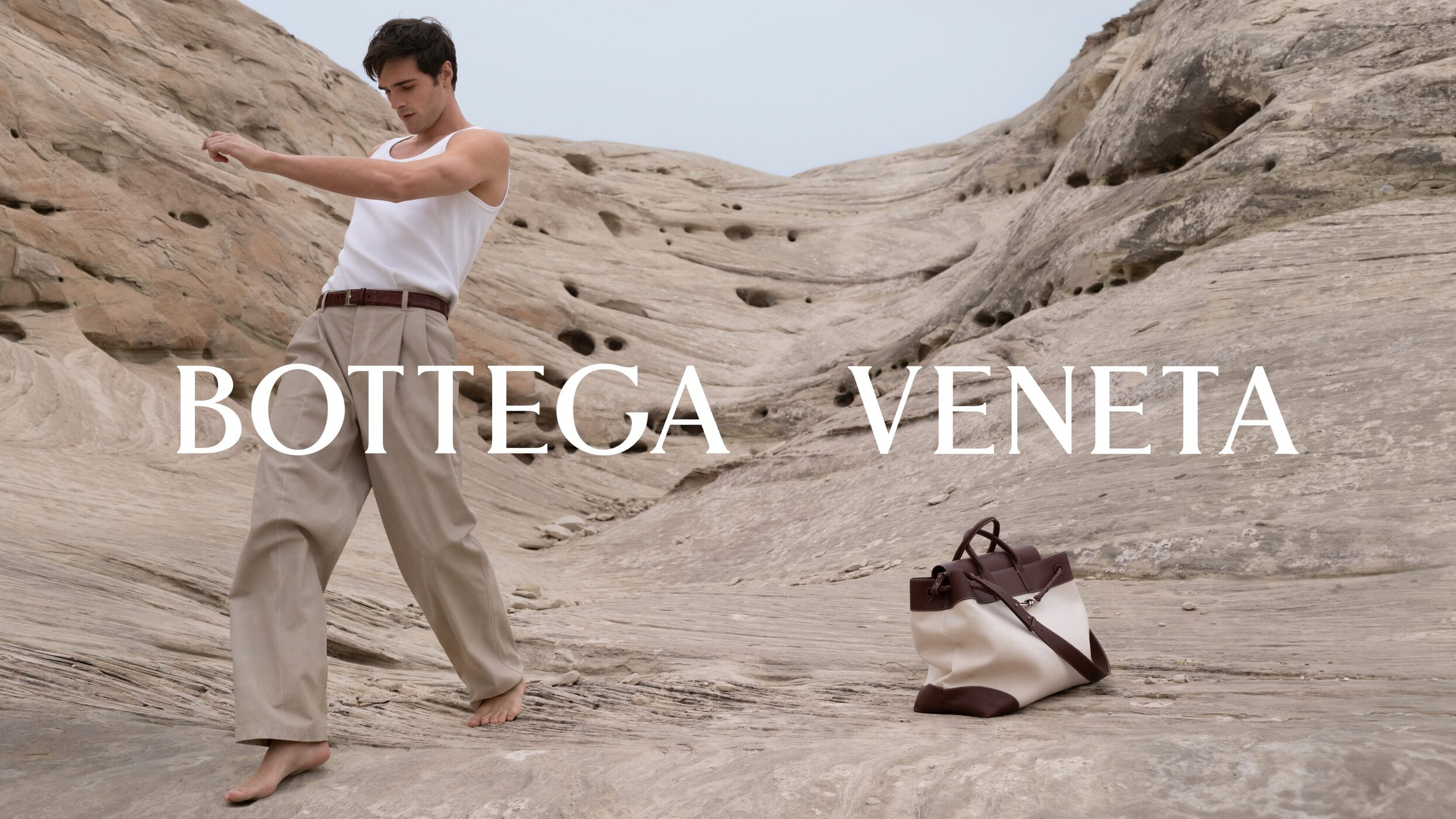 Jacob Elordi wearing a white tank top and beige trousers standing barefoot in a rocky landscape next to a canvas handbag